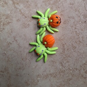 May include: Two green and purple spiders with orange pumpkin bodies. The pumpkins have carved jack-o'-lantern faces.