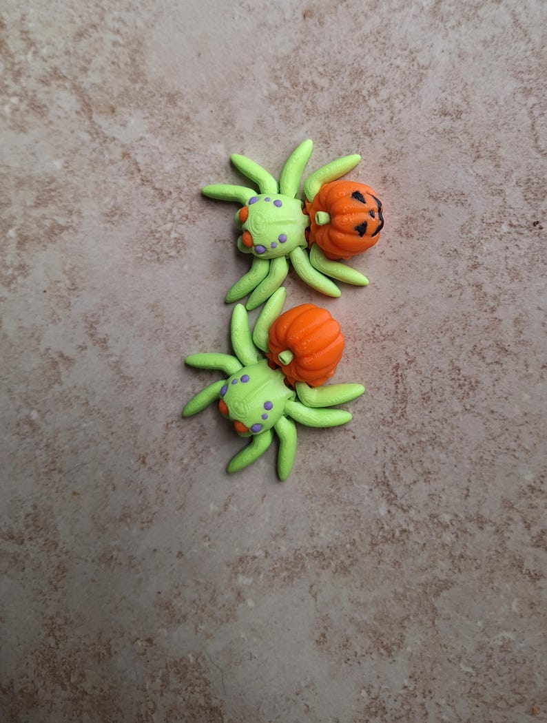 May include: Two green and purple spiders with orange pumpkin bodies. The pumpkins have carved jack-o'-lantern faces.