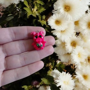 May include: A small, handmade pink dragon charm with black eyes and a red strawberry. The dragon has small horns and a loop for attaching to a necklace or bracelet. The charm is held in front of a background of white flowers.