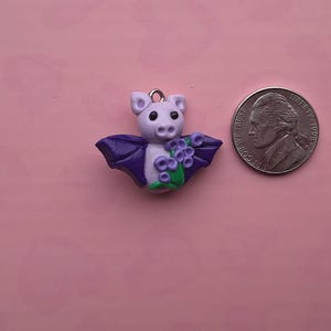May include: A handcrafted bat charm made of polymer clay. The bat has a light purple body, dark purple wings, and holds a bouquet of purple flowers. A nickel coin is next to the charm for size reference.