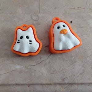 K&ouml;nnte beinhalten: Zwei Halloween-Ornamente. Eines ist ein wei&szlig;es, katzenf&ouml;rmiges Design mit schwarzen Augen und Schnurrhaaren, orange umrandet. Das andere ist ein wei&szlig;er Geist, der einen K&uuml;rbis h&auml;lt, ebenfalls orange umrandet. Beide haben eine Schlaufe zum Aufh&auml;ngen.