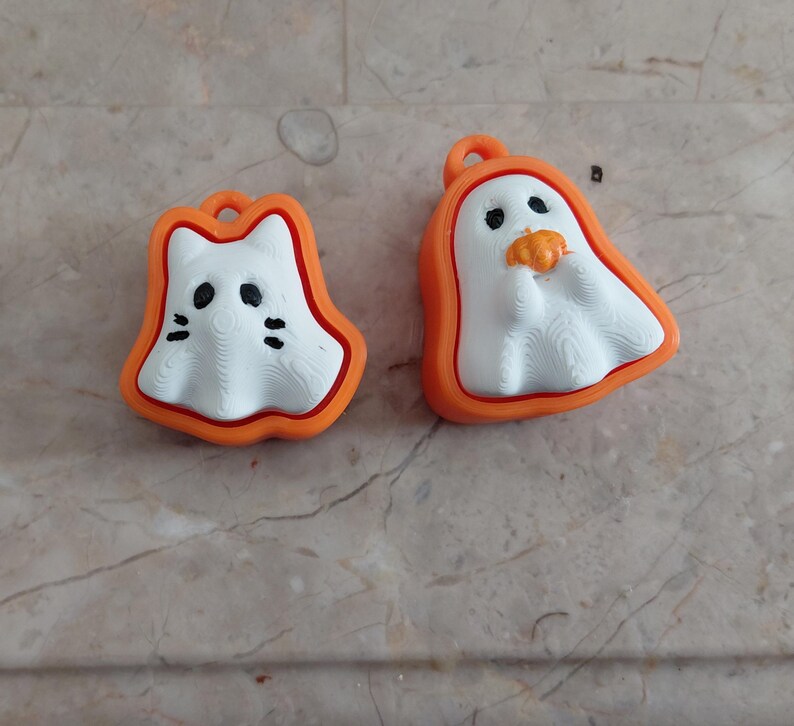 K&ouml;nnte beinhalten: Zwei Halloween-Ornamente. Eines ist ein wei&szlig;es, katzenf&ouml;rmiges Design mit schwarzen Augen und Schnurrhaaren, orange umrandet. Das andere ist ein wei&szlig;er Geist, der einen K&uuml;rbis h&auml;lt, ebenfalls orange umrandet. Beide haben eine Schlaufe zum Aufh&auml;ngen.