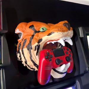 May include: A red and black video game controller is held in the mouth of a tiger head wall mount. The tiger head is orange and black with white teeth.