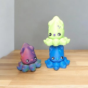 Puede incluir: Tres peluches de calamar de colores apilados uno encima del otro. El calamar de arriba es verde, el calamar del medio es azul y el calamar de abajo es morado.