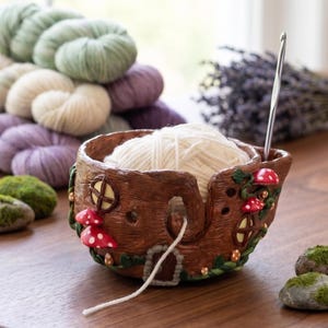 May include: A brown ceramic yarn bowl shaped like a tree trunk with red mushroom accents and a small door. The bowl holds a ball of white yarn, with a crochet hook inserted. Green, white, and purple yarn skeins are in the background.
