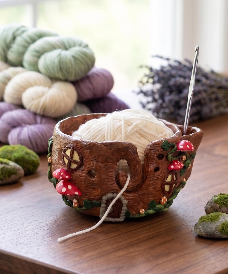 May include: A brown ceramic yarn bowl shaped like a tree trunk with red mushroom accents and a small door. The bowl holds a ball of white yarn, with a crochet hook inserted. Green, white, and purple yarn skeins are in the background.