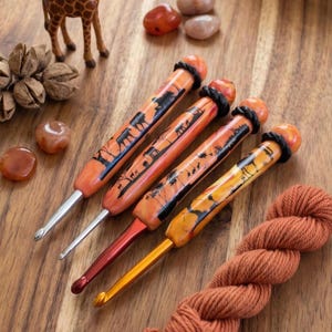 May include: Four crochet hooks with orange and yellow marbled handles featuring black silhouettes of African wildlife, including elephants and giraffes. Each hook has a black ring accent near the top and a metallic tip in silver or gold.