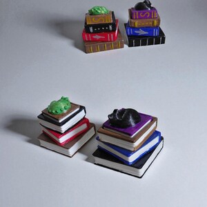 K&ouml;nnte beinhalten: Miniaturstapel von B&uuml;chern in verschiedenen Farben, jeweils gekr&ouml;nt von einer kleinen Figur. Die Figuren umfassen ein gr&uuml;nes Blatt und eine schwarze Katze. Die B&uuml;cher sind in Braun-, Rot-, Schwarz-, Blau- und Lilat&ouml;nen bemalt.