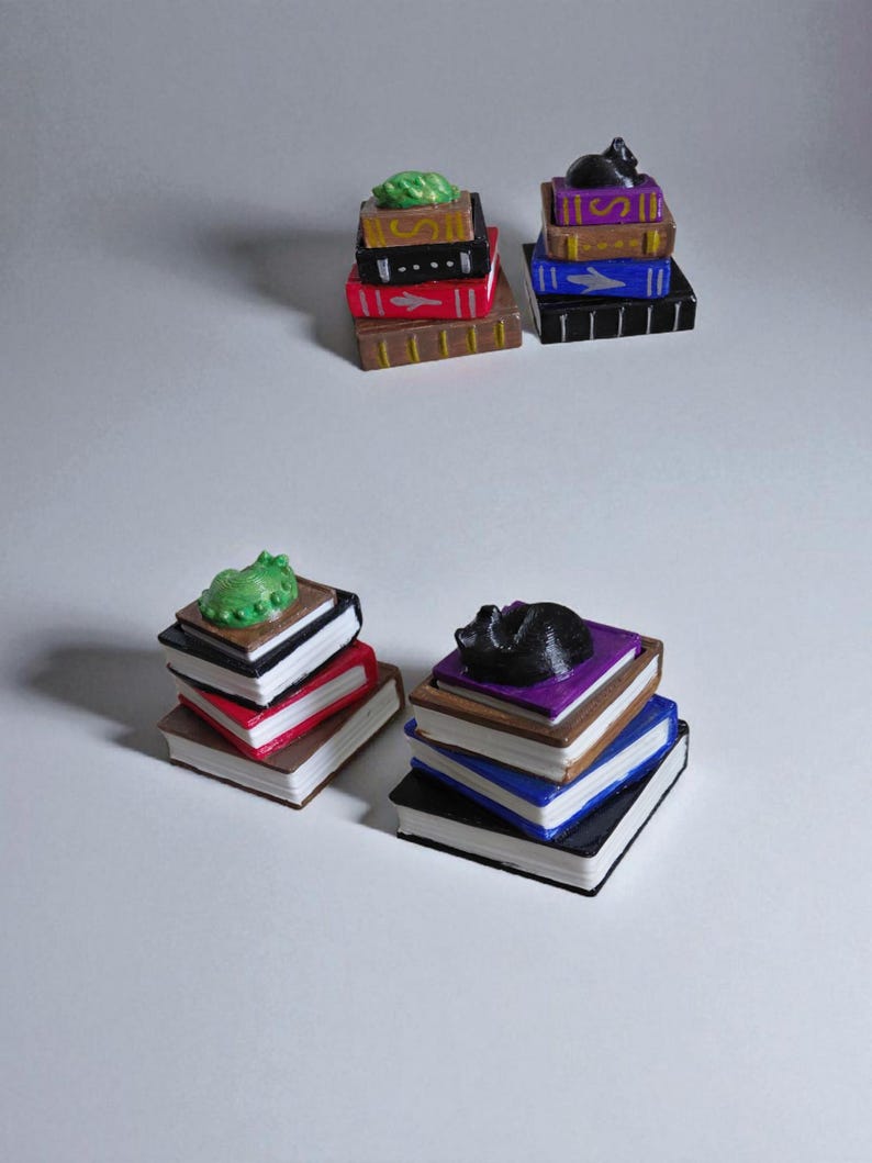 K&ouml;nnte beinhalten: Miniaturstapel von B&uuml;chern in verschiedenen Farben, jeweils gekr&ouml;nt von einer kleinen Figur. Die Figuren umfassen ein gr&uuml;nes Blatt und eine schwarze Katze. Die B&uuml;cher sind in Braun-, Rot-, Schwarz-, Blau- und Lilat&ouml;nen bemalt.