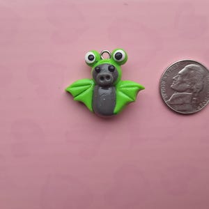 May include: A handmade polymer clay bat charm with a green body, gray belly, and large, round eyes. The charm is next to a US nickel coin for size comparison. The background is a solid pink color.