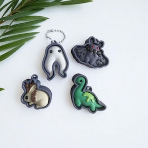 Op de afbeelding: Vier grillige sleutelhangers in de vorm van een spook, vleermuis, konijn en dinosaurus. De bedels hebben een donkergrijze rand met de binnenontwerpen in wit, bruin en groen. De spookbedel heeft een zilveren ketting.