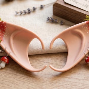 Puede incluir: Un par de orejas de elfo rosas con detalles rojos y blancos, musgo verde y dijes de mariposa plateados. Cada oreja tiene un dije de hongo colgante. Para cosplay o disfraces.
