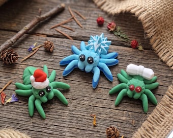 May include: Three small spider figurines made of clay. One green spider wears a red and white Santa hat. A central blue spider features a white snowflake pattern on its back. A third green spider has red eyes and a white rectangular object on its back.