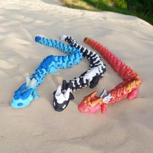 Puede incluir: Tres juguetes de dragón segmentados y flexibles. Los dragones son azul, negro y blanco, y rojo con detalles dorados. Los dragones están colocados sobre una superficie arenosa.