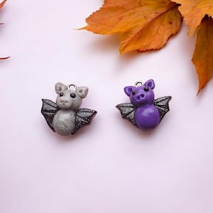 May include: Two small bat charms made of polymer clay. One is gray with black wings and the other is purple with black wings.