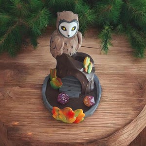 以下が含まれることがあります： 茶色と白のフクロウの置物が、灰色の土台のある木の切り株の上に置かれています。土台にはサイコロを入れるためのスロットがあり、茶色とオレンジの葉のデザインが施されています。土台には、紫、オレンジ、赤のサイコロが3つあります。