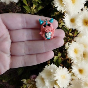 Op de afbeelding: Een handgemaakt roze drakenbeeldje met blauwe hoorns en accenten, met een zeeschelp. De draak heeft zwarte ogen en een zilveren lus om op te hangen. Het beeldje is een grillige, fantasie-thema charme.