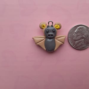 May include: A handmade bat charm made of polymer clay. The bat has a gray body, tan wings, and yellow accents on its ears. A silver-colored metal loop is attached to the top for hanging. The charm is next to a US nickel coin.