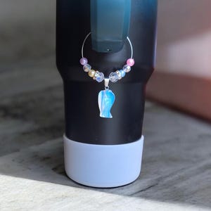 May include: A silver wine charm with a blue and white angel charm and iridescent beads. The charm is hanging from a black and blue tumbler.