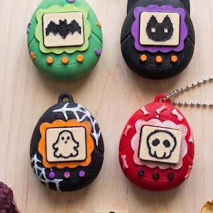 Könnte beinhalten: Vier eiförmige digitale Haustier-Schlüsselanhänger in Grün, Schwarz und Rot mit Halloween-Motiven wie Fledermaus, Katze, Geist und Totenkopf auf den Frontplatten. Darunter befinden sich drei quadratische Plättchen mit Spinne, Kürbis und Hexenhut.