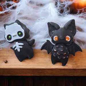 May include: Two Halloween-themed figurines, a black and white skeleton and a black bat with orange eyes, are displayed on a wooden surface.