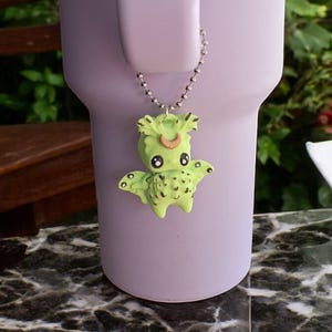May include: A green dragon charm with black eyes and a brown horn hangs from a silver chain. The charm is attached to a purple mug.