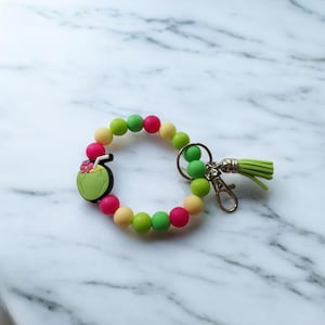 Peut inclure: Un bracelet coloré en perles avec un charme en forme de noix de coco et un porte-clés. Le bracelet est composé de perles vertes, jaunes et roses. Le charme est une noix de coco de dessin animé avec une paille et une fleur.