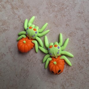 May include: Two green and purple spiders with orange pumpkins. The pumpkins have jack-o'-lantern faces.