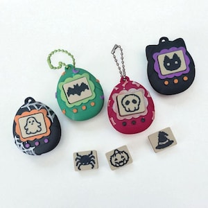 Op de afbeelding: Een verzameling Halloween-thema digitale huisdier speelgoed in verschillende kleuren. Elk speelgoed heeft een ander griezelig icoon, zoals een spook, vleermuis of schedel. Kleine, vierkante bedels met Halloween-ontwerpen zijn ook aanwezig.