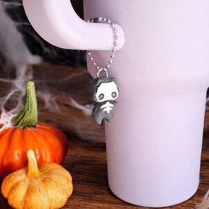Puede incluir: Una pequeña figura de esqueleto gris y blanco con ojos verdes cuelga de una cadena plateada unida a una taza morada. La taza está sobre una superficie de madera con dos pequeñas calabazas.