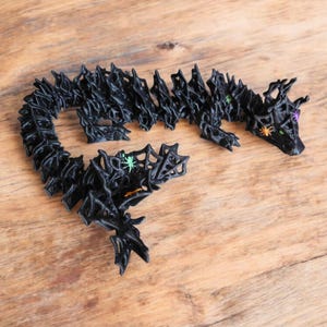 May include: A black, articulated dragon figurine with a textured, spiky design. The dragon has green and purple eyes, and is decorated with small spider accents. The figurine is displayed on a wooden surface.
