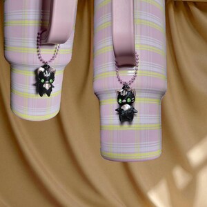 Może przedstawiać: Dwa różowe kubki z wzorem w kratę i jasnoróżowym uchwytem. Każdy kubek ma czarny urok kota z zielonymi oczami wiszący na łańcuszku z koralików. Tło jest jasnobrązowe.