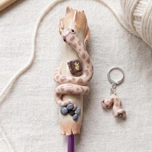 Puede incluir: Un ganchillo con un mango de color tostado claro esculpido en forma de serpiente que se enrosca alrededor del eje. La serpiente tiene manchas marrones y ojos oscuros. Un pequeño libro marrón con un símbolo dorado y piedras grises decoran el mango. Llavero a juego.