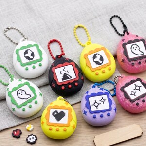 May include: A collection of eight egg-shaped digital pet keychains in various colors including white, black, yellow, pink, and purple. Each device features a small screen displaying pixelated icons, three circular buttons, and a metal ball chain.