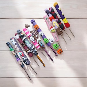 Op de afbeelding: Een verzameling van 14 kleurrijke haaknaalden met Halloween-thema's. De handvatten hebben verschillende karakters en ontwerpen, waaronder pompoenen, geesten en een peperkoekhuis.