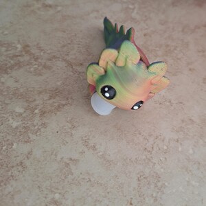 Pu&ograve; includere: Una piccola statuina di axolotl di cartone animato stampata in 3D, colorata, con grandi occhi. L'axolotl &egrave; verde, giallo e arancione con una base bianca.