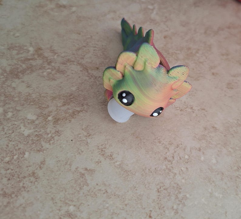 Pu&ograve; includere: Una piccola statuina di axolotl di cartone animato stampata in 3D, colorata, con grandi occhi. L'axolotl &egrave; verde, giallo e arancione con una base bianca.