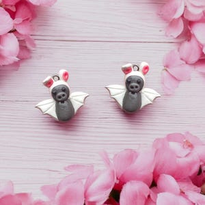 May include: Two small gray and white bat charms with pink bunny ears. The charms are on a pink wooden background with pink flowers.