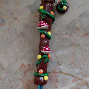 May include: A handcrafted pencil with a brown clay shaft, adorned with green vines, red and white spotted mushrooms, and yellow and orange spheres. A matching charm with a mushroom and green leaves hangs from the top.
