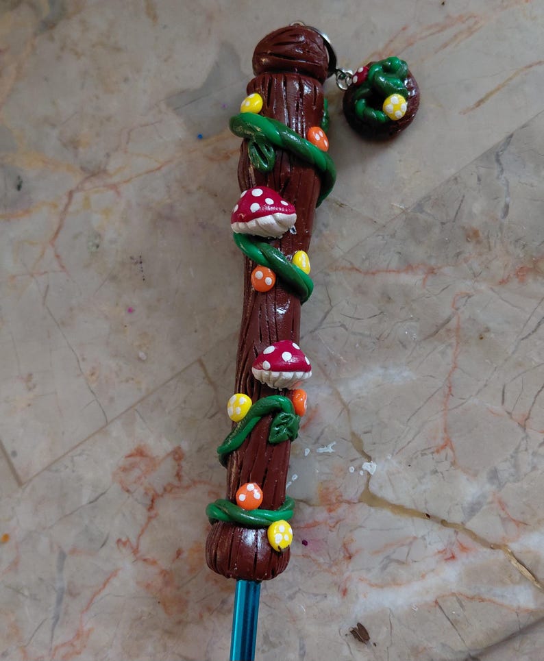 May include: A handcrafted pencil with a brown clay shaft, adorned with green vines, red and white spotted mushrooms, and yellow and orange spheres. A matching charm with a mushroom and green leaves hangs from the top.