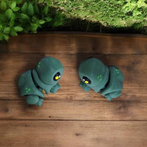 Puede incluir: Dos pequeñas figuritas verdes y negras con cuerpos segmentados y ojos amarillos. Las figuritas están sobre una superficie de madera.
