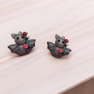 May include: A pair of gray bat earrings with pink and red flower accents. The bats have a cute, cartoon-like appearance.