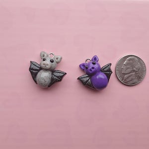 May include: Two small bat-shaped charms. One is gray with black glitter wings, the other is purple with black glitter wings. A US nickel coin is next to the charms for size comparison. The background is a solid pink color.