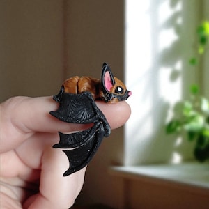 Könnte beinhalten: Eine Miniatur-Fledermausfigur mit braunem Fell, schwarzen Flügeln und rosa Innenohren. Die Fledermaus liegt, die Flügel ausgebreitet. Die Figur ist klein und detailliert, mit einem niedlichen Ausdruck.
