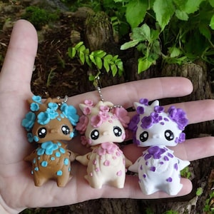 May include: Three handmade fantasy creature figurines in brown, cream, and white, each adorned with floral accents in blue, pink, and purple. The figurines have large black eyes and are held in a hand.