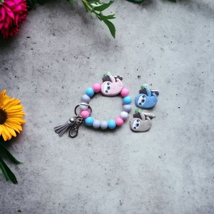 May include: A gray, blue, and pink silicone bracelet with a silver key chain and three sloth charms. The charms are gray, blue, and pink.