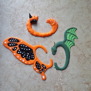 以下が含まれることがあります： オレンジと黒の3Dプリントされた動物の置物3つ。丸まったドラゴン、蝶、丸まったキツネ