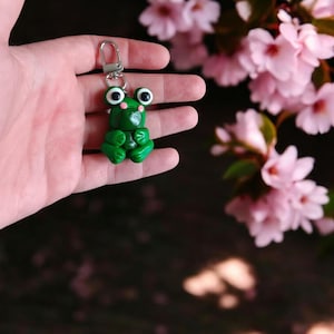 May include: A green frog keychain with large, white eyes and a silver ring. The frog is sitting on a hand with pink flowers in the background.