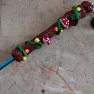 May include: A handcrafted crochet hook with a brown wooden handle, adorned with green vines, red and white mushroom accents, and yellow and orange dots. A matching charm hangs from the end. The hook itself is a vibrant blue.