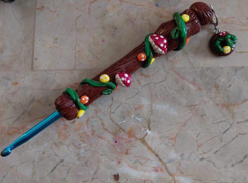 May include: A handcrafted crochet hook with a brown wooden handle, adorned with green vines, red and white mushroom accents, and yellow and orange dots. A matching charm hangs from the end. The hook itself is a vibrant blue.